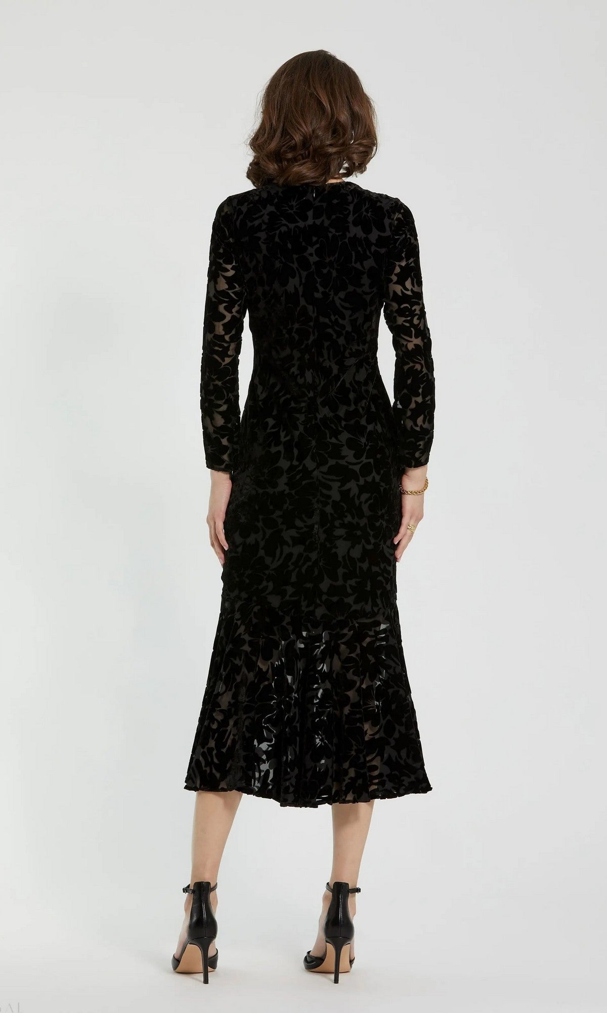 A woman stands with her back to the camera, wearing the Mac Duggal 81129 midi party dress in black burnout velvet with long sleeves and paired with black high-heeled shoes, set against a plain white background.